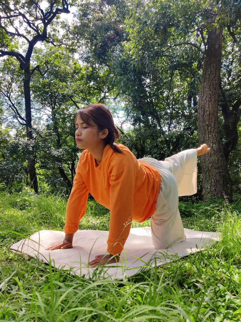 K&ouml;nnte beinhalten: Eine Frau in einem orangefarbenen Langarmshirt und einer wei&szlig;en Hose f&uuml;hrt eine Yoga-Pose auf einer wei&szlig;en Matte in einem grasbewachsenen Bereich mit B&auml;umen im Hintergrund aus.