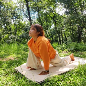 K&ouml;nnte beinhalten: Eine Person in einem orangenen Shirt und einer wei&szlig;en Hose macht eine Yoga-Pose auf einer wei&szlig;en Matte in einem grasbewachsenen Bereich. Die Person schaut nach oben und hat ihre Arme vor sich ausgestreckt.