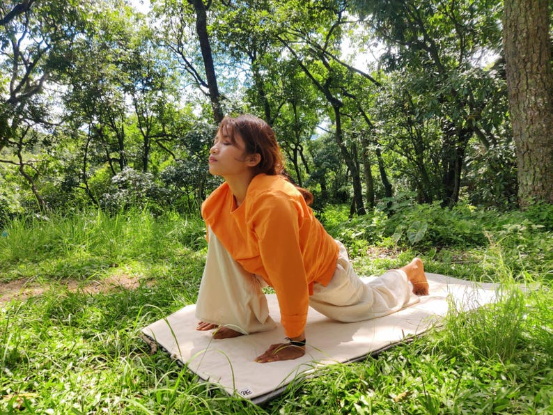 K&ouml;nnte beinhalten: Eine Person in einem orangenen Shirt und einer wei&szlig;en Hose macht eine Yoga-Pose auf einer wei&szlig;en Matte in einem grasbewachsenen Bereich. Die Person schaut nach oben und hat ihre Arme vor sich ausgestreckt.