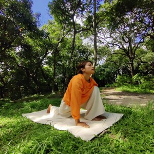 K&ouml;nnte beinhalten: Eine Frau in einem orangenen Shirt und einer wei&szlig;en Hose macht eine Yoga-Pose auf einer wei&szlig;en Matte in einem grasbewachsenen Bereich mit B&auml;umen im Hintergrund.