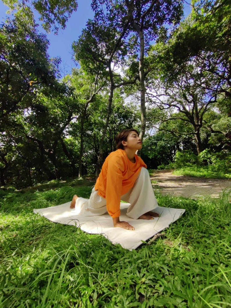 K&ouml;nnte beinhalten: Eine Frau in einem orangenen Shirt und einer wei&szlig;en Hose macht eine Yoga-Pose auf einer wei&szlig;en Matte in einem grasbewachsenen Bereich mit B&auml;umen im Hintergrund.