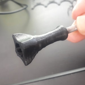 Może przedstawiać: Czarny plastikowy uchwyt do kamery z gwintowaną śrubą metalową. Uchwyt ma trójkątny kształt na końcu.