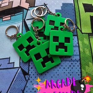 May include: A collection of green square keyrings, each featuring a black pixelated face design. The keyrings are attached to silver metal rings. The background has a cartoon graphic and the text "ARCADE GRENADE".