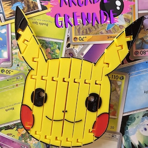 Puede incluir: Un juguete de rompecabezas amarillo con forma de Pikachu con ojos negros, mejillas rojas y orejas con puntas negras. El juguete está hecho de piezas entrelazadas. El fondo presenta varias cartas coleccionables de Pokémon, con el texto "Arcade Grenade".