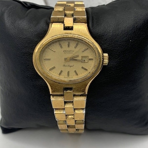 May include: A gold-toned Zenith automatic wristwatch with a gold-tone metal band. The watch face is round and has a gold-tone bezel. The watch face has black Roman numeral hour markers, black hands, and a date window at the 3 o'clock position. The watch face also has the text "Zenith Automatic" and "Port Royal" printed on it.