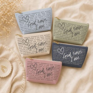 May include: Five rectangular zippered pouches in light blue, sage green, cream, navy blue, and dusty rose. Each pouch features a gold zipper and a printed design with a heart outline and the text "God says I am" followed by various inspirational words.
