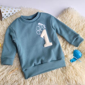 Könnte beinhalten: Ein blaues Sweatshirt mit Waffelstruktur und langen Ärmeln. Es hat den Namen "Moritz" über einer weißen Zahl "1" und einem Ballondesign. Das Kleidungsstück ist für den ersten Geburtstag eines Kindes gedacht.