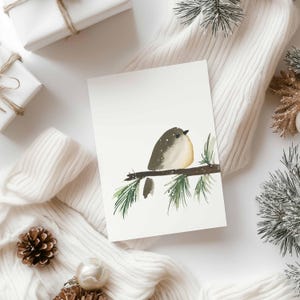 Può includere: Un biglietto di auguri ad acquerello con un uccellino appollaiato su un ramo con aghi verdi. L'uccello ha il dorso grigio, il petto giallo chiaro e un occhio scuro. Il biglietto è circondato da regali bianchi, un maglione bianco e pigne.
