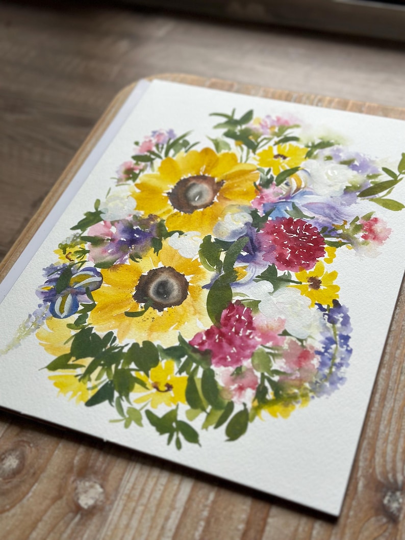 May include: A watercolor painting on white paper featuring a vibrant floral arrangement. The composition includes large yellow sunflowers with dark centers, interspersed with smaller pink, purple, and white blossoms, surrounded by green foliage.