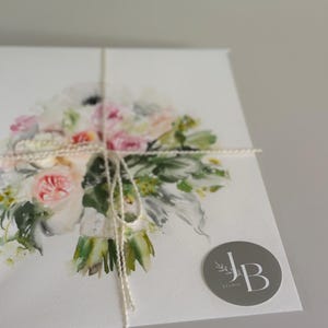 May include: A white card featuring a watercolor illustration of a floral bouquet with pink, white, and green tones. The card is tied with thin white twine and includes a circular gray sticker in the bottom right corner with the text JB STUDIO.