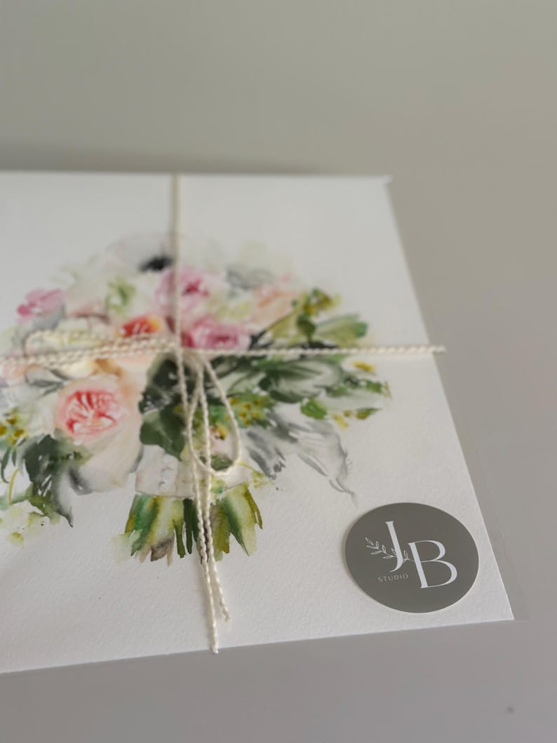 May include: A white card featuring a watercolor illustration of a floral bouquet with pink, white, and green tones. The card is tied with thin white twine and includes a circular gray sticker in the bottom right corner with the text JB STUDIO.