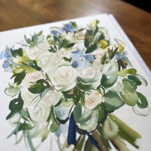 May include: A watercolor painting on white paper depicting a floral bouquet with white roses, green foliage, and light blue accents. The arrangement is tied with a dark blue ribbon at the base, featuring loose, expressive brushstrokes throughout.