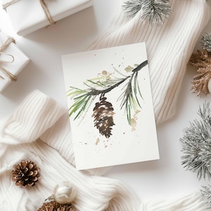 Może przedstawiać: Akwarela szyszki i gałęzi z zielonymi igłami. Grafika znajduje się na białej karcie, otoczona białym swetrem, zapakowanymi prezentami i dekoracyjnymi szyszkami. Ogólnym motywem jest zima i natura.