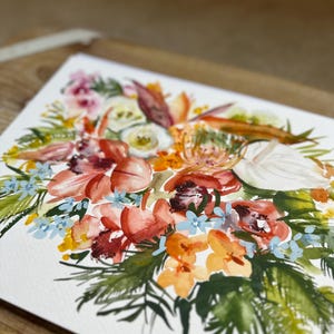 May include: A watercolor painting on white paper featuring a vibrant floral arrangement. The composition includes large coral and orange blossoms, delicate light blue flowers, white petals, and various green foliage, all rendered with fluid brushstrokes.