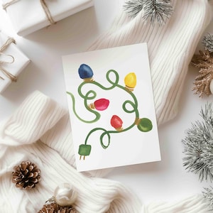 May include: A white greeting card with a watercolour illustration of Christmas lights in blue, yellow, red, and green. The card is set against a white sweater, wrapped gifts, pine cones, and baubles, creating a festive holiday theme.