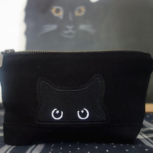 May include: A black zippered pouch featuring an embroidered patch of a cat head with white eyes peeking over the top edge. The pouch is made of a solid black fabric and includes a metal zipper pull on the left side for opening and closing.