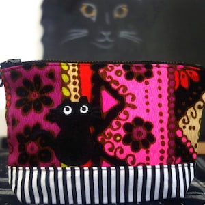 May include: A rectangular zippered pouch featuring a vibrant pattern of pink, magenta, and brown floral and geometric shapes. A black cat applique with white eyes is centered on the front. The bottom section has a black and white striped border.