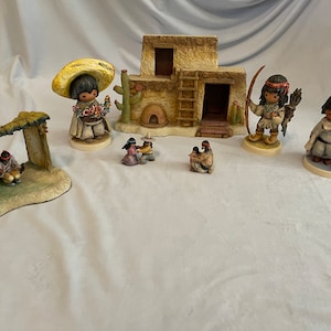 May include: A set of Native American figurines depicting a family, a hunter, and a woman holding flowers. The figurines are made of painted ceramic and are displayed on a white background. The set includes a small adobe house with a ladder and a cactus.