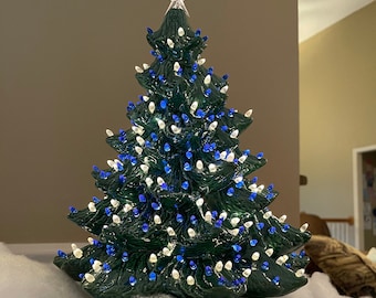 May include: A green ceramic Christmas tree with white and blue lights. The tree is decorated with a star on top and is sitting on a white fluffy surface.