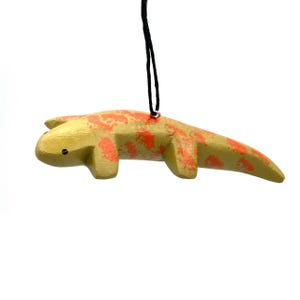 May include: A wooden ornament shaped like a lizard with orange and yellow paint. The ornament has a black string for hanging.