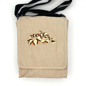 May include: Beige crossbody bag with a black strap. The bag features an embroidered snake design in shades of brown, tan, and cream. The bag has a flap closure and a rectangular shape, suitable for carrying essentials.