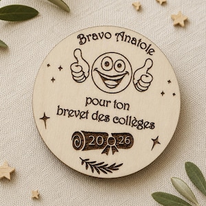 Peut inclure: Une plaque circulaire en bois clair gravée d'un emoji souriant faisant deux pouces levés. Le texte indique Bravo Anatole pour ton brevet des collèges, avec un motif de diplôme, l'année 2026 et de petites étoiles décoratives.