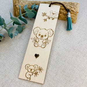 May include: A wooden bookmark with engraved koala bear designs. It has a brown leather cord and a teal tassel. The koala illustrations include a sitting koala, a koala on a branch, and a heart-shaped cutout.
