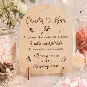 Peut inclure: Panneau en bois "Candy Bar" avec un cœur découpé et du texte gravé. Le panneau comprend des phrases comme "Faites vous plaisir" et "Servez-vous", ainsi que des illustrations de bonbons. Divers bonbons et friandises entourent le panneau.