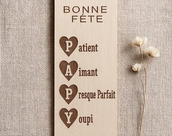 May include: Wooden Father's Day gift tag with the words "Bonne Fête" and the words "Patient, Aimant, Presque Parfait, Youpi" in a heart shape. A brown leather cord and a small wooden bead are attached to the tag. A small sprig of dried flowers is to the right.