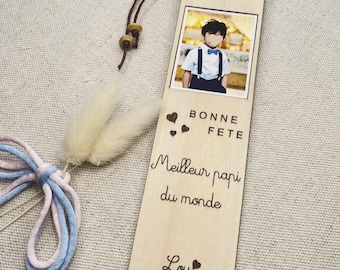 May include: Wooden bookmark with a photo of a child wearing a bow tie and suspenders. The bookmark has the text "BONNE FETE Meilleur papi du monde Lou" and is adorned with a brown cord and decorative elements.