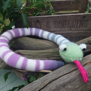 Peut inclure: Un jouet serpent tricot&eacute; avec un corps ray&eacute; violet, blanc et bleu et une t&ecirc;te verte avec une langue rose. Le serpent est allong&eacute; sur une b&ucirc;che de bois.