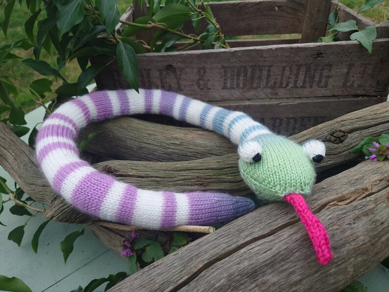 Peut inclure: Un jouet serpent tricot&eacute; avec un corps ray&eacute; violet, blanc et bleu et une t&ecirc;te verte avec une langue rose. Le serpent est allong&eacute; sur une b&ucirc;che de bois.