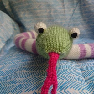 Peut inclure: Un jouet en forme de grenouille verte tricot&eacute;e avec un corps ray&eacute; blanc et violet et une langue rose. La grenouille est pos&eacute;e sur une surface &agrave; motifs bleus et blancs.