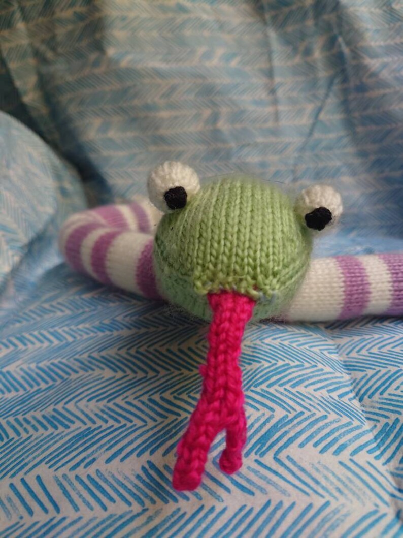 Peut inclure: Un jouet en forme de grenouille verte tricot&eacute;e avec un corps ray&eacute; blanc et violet et une langue rose. La grenouille est pos&eacute;e sur une surface &agrave; motifs bleus et blancs.
