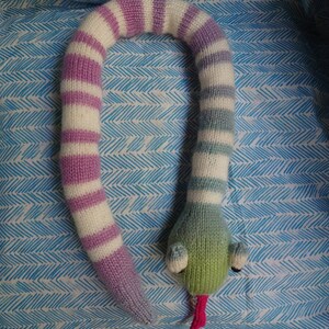 Peut inclure: Un jouet serpent tricot&eacute; avec des rayures roses et blanches d'un c&ocirc;t&eacute; et des rayures bleues et blanches de l'autre. Le serpent a une t&ecirc;te verte avec des yeux roses et une langue rose.