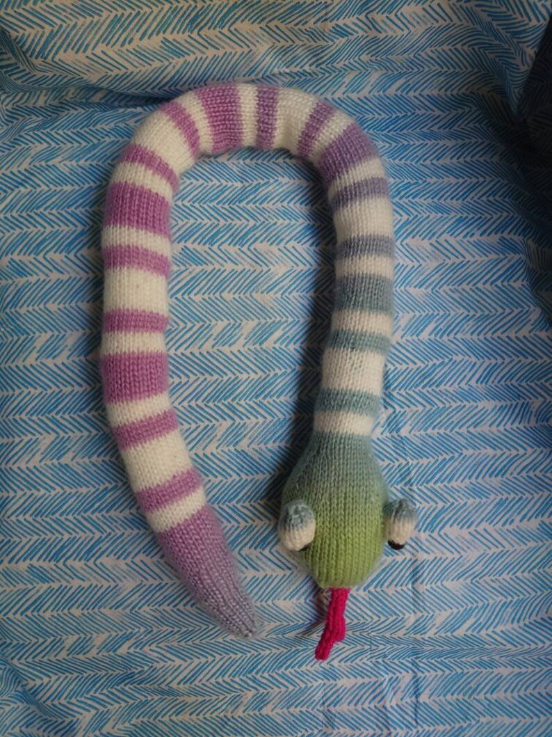 Peut inclure: Un jouet serpent tricot&eacute; avec des rayures roses et blanches d'un c&ocirc;t&eacute; et des rayures bleues et blanches de l'autre. Le serpent a une t&ecirc;te verte avec des yeux roses et une langue rose.