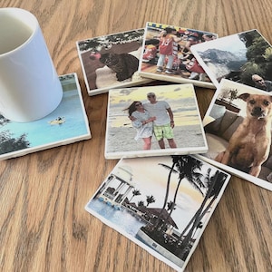 May include: A white ceramic mug rests on a square coaster featuring a scenic image. Several other coasters with various photo prints are scattered around, including a cat, people, a dog, and a tropical scene. The coasters are square and appear to be made of stone.