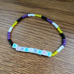 May include: A beaded bracelet with black, white, yellow, and purple beads. The bracelet spells out "NONBINARY" in colorful letters.