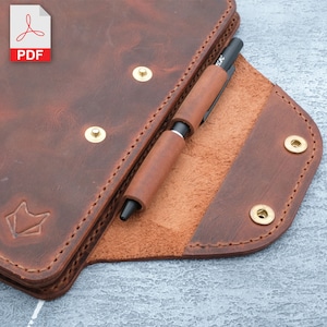 May include: A brown leather notebook cover with a pen holder and brass rivets. The cover is open to reveal a brown leather interior.