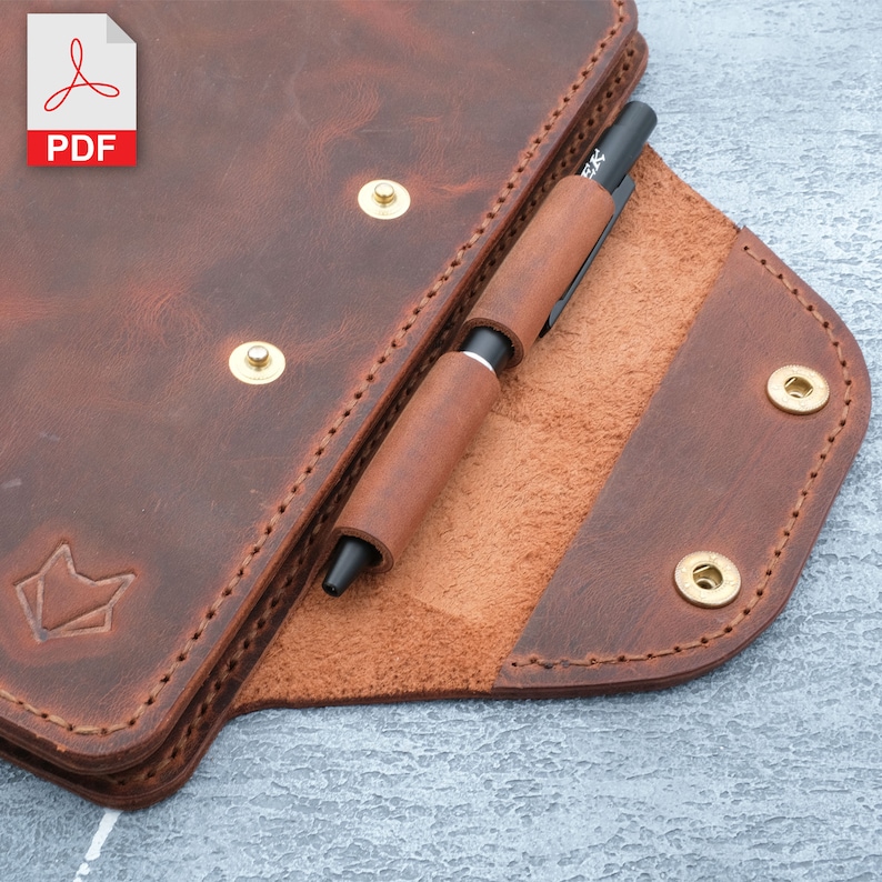 May include: A brown leather notebook cover with a pen holder and brass rivets. The cover is open to reveal a brown leather interior.