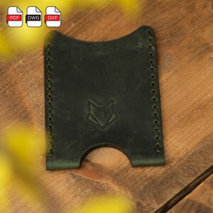 May include: A dark green leather card holder with a fox logo embossed on the front. The card holder has a stitched edge and a slot for cards.