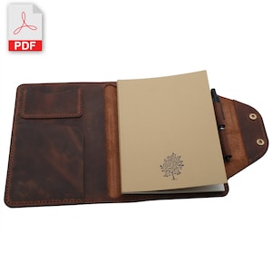 May include: A brown leather notebook cover with a pen holder and a pocket for a small notebook. The notebook cover is open to reveal a blank notebook with a tree design printed on the front.