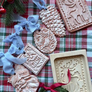 May include: A collection of terracotta-colored Christmas ornaments with intricate designs, including a pinecone, a bird, and a reindeer. Each ornament is attached to a light blue ribbon. The ornaments are arranged on a red and green plaid background.
