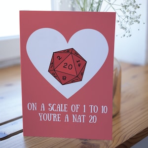 May include: A red and white card with a heart shape and a red twenty-sided die inside. The text on the card reads "On a scale of 1 to 10 you're a Nat 20".