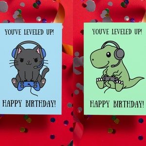 May include: Two greeting cards with a blue and green background. The cards feature a black cat and a green dinosaur wearing headphones and holding a video game controller. The text on the cards reads "You've leveled up! Happy Birthday!"