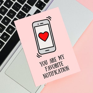 May include: A pink card with a black outline of a smartphone with a red heart on the screen. The text on the card reads "You are my favorite notification."