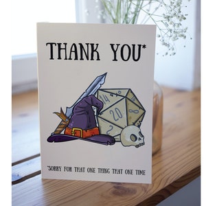 May include: A thank you card with a cartoon illustration of a wizard's hat, a dagger, a 20-sided die, and a skull. The text on the card reads "THANK YOU*" and "SORRY FOR THAT ONE THING THAT ONE TIME".