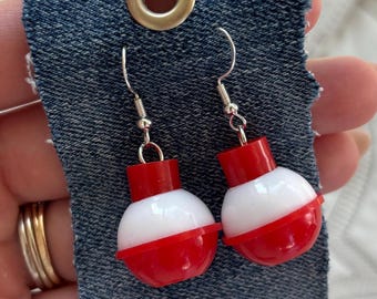 May include: A pair of dangle earrings featuring spherical fishing bobber charms in red and white plastic. The earrings are attached to a rectangular piece of dark blue denim fabric with a silver metal grommet at the top for display purposes.