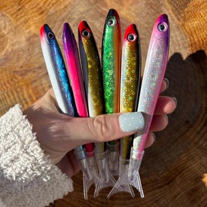 May include: A collection of six colorful fish-shaped pens. The pens feature a variety of colors, including blue, pink, green, and silver, with red accents. Each pen has a clear plastic tail and a silver pen tip. The pens are held together.