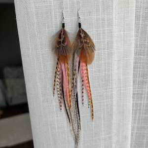 May include: A pair of feather earrings with a mix of brown, pink, and black feathers. The earrings have silver hooks and black accents. The feathers are arranged in a layered design, creating a flowing, bohemian look.